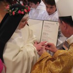 Photo en plongée sur l'évêque passant une alliance au doigt d'une sœur revêtue du voile noir et d'une couronne de roses.
