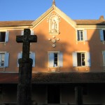 Vue du cloître, la façade rose du monastère est caressée par le soleil d'un matin d'été. A son sommet, la Vierge veille sur le cloître. En premier plan une croix de pierre habillée de lichens ocres-jaunes.