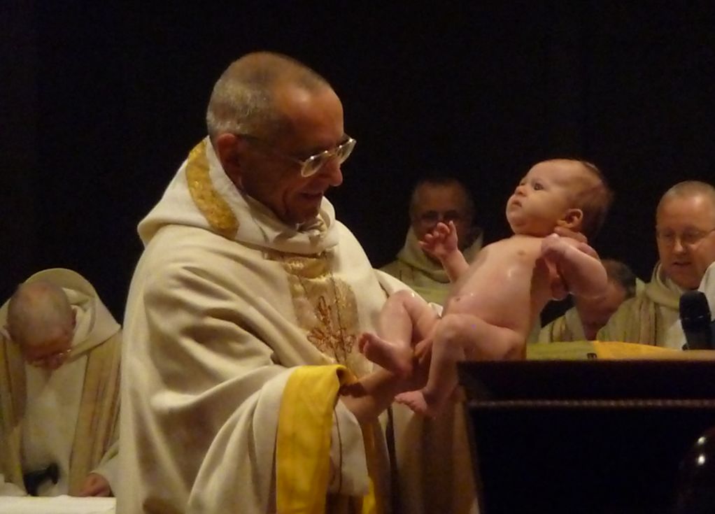 Le père abbé tient un bébé dans ses mains, à la sortie de la cuve baptismale.